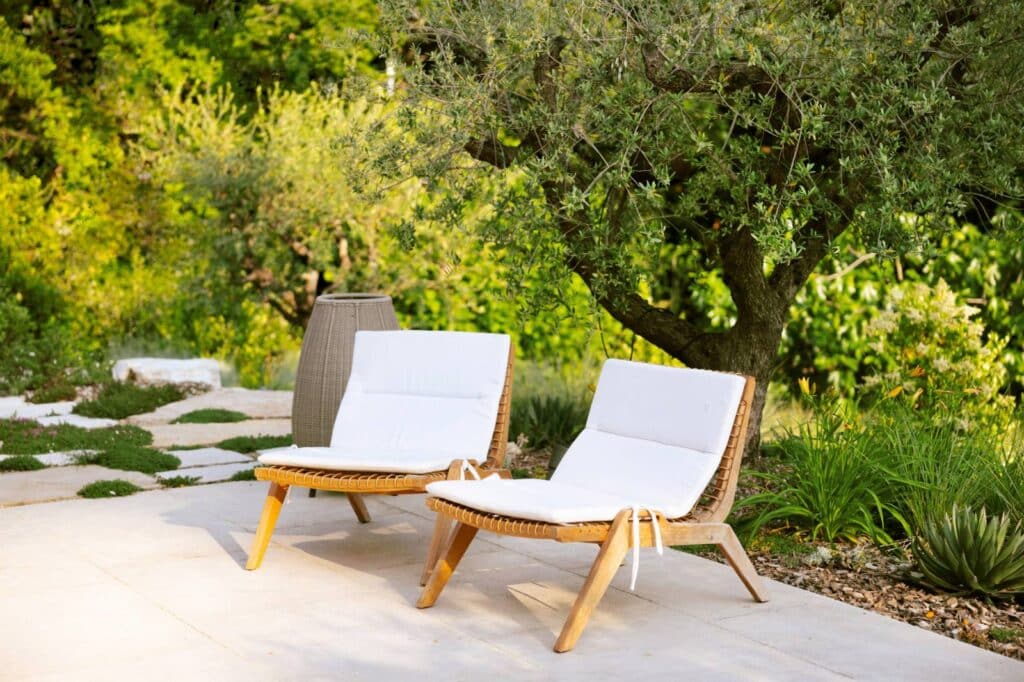 Deux chaises longues en bois avec coussins blancs, sur une terrasse claire, sous un olivier, entourées d'un jardin verdoyant.