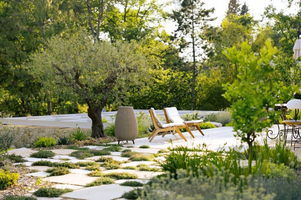 Cheminement minéral en pierre naturelle, structurant les circulations au cœur d’un jardin immersif à Châteauneuf-de-Grasse. Terrasse extérieure moderne avec dalles de pierre et plantes couvre-sol, chaises longues en bois et grand lampadaire tressé près d'un olivier.