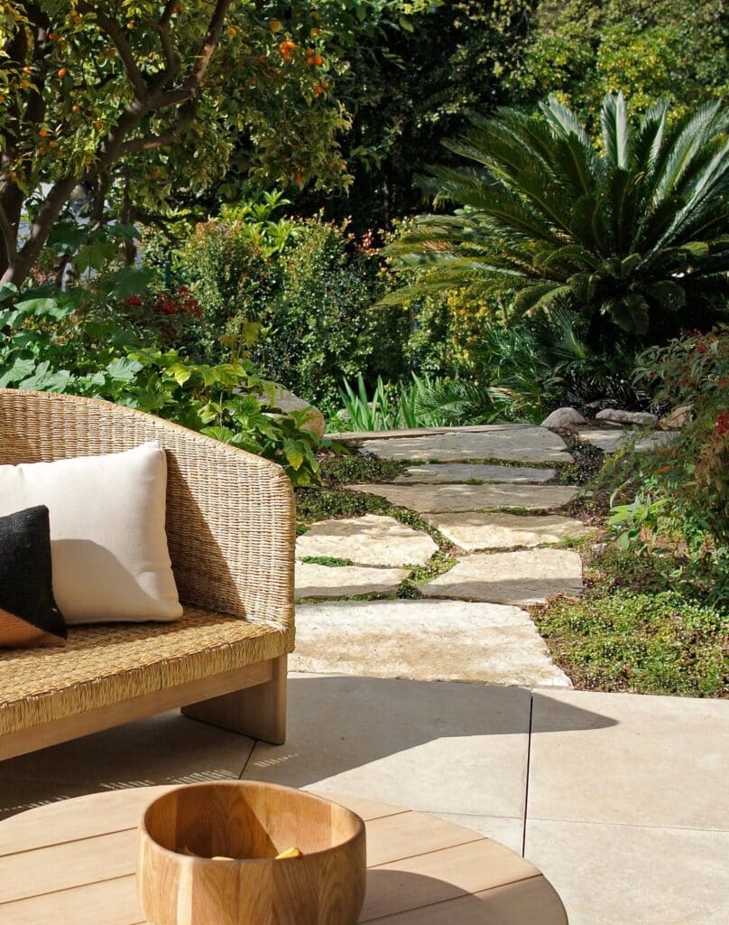 Patio avec canapé en osier et coussins. Chemin de pierre traversant un jardin luxuriant avec palmier, arbre à agrumes et plantes vertes.