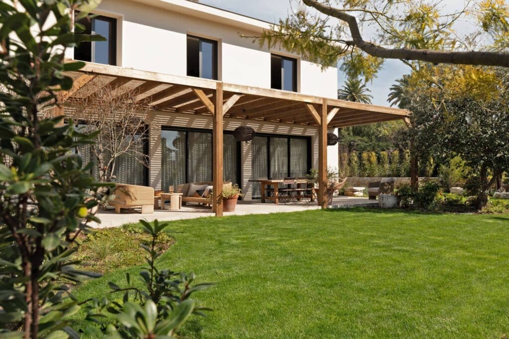 Maison moderne blanche avec grande pergola en bois et terrasse aménagée, salon extérieur, salle à manger et pelouse verte.