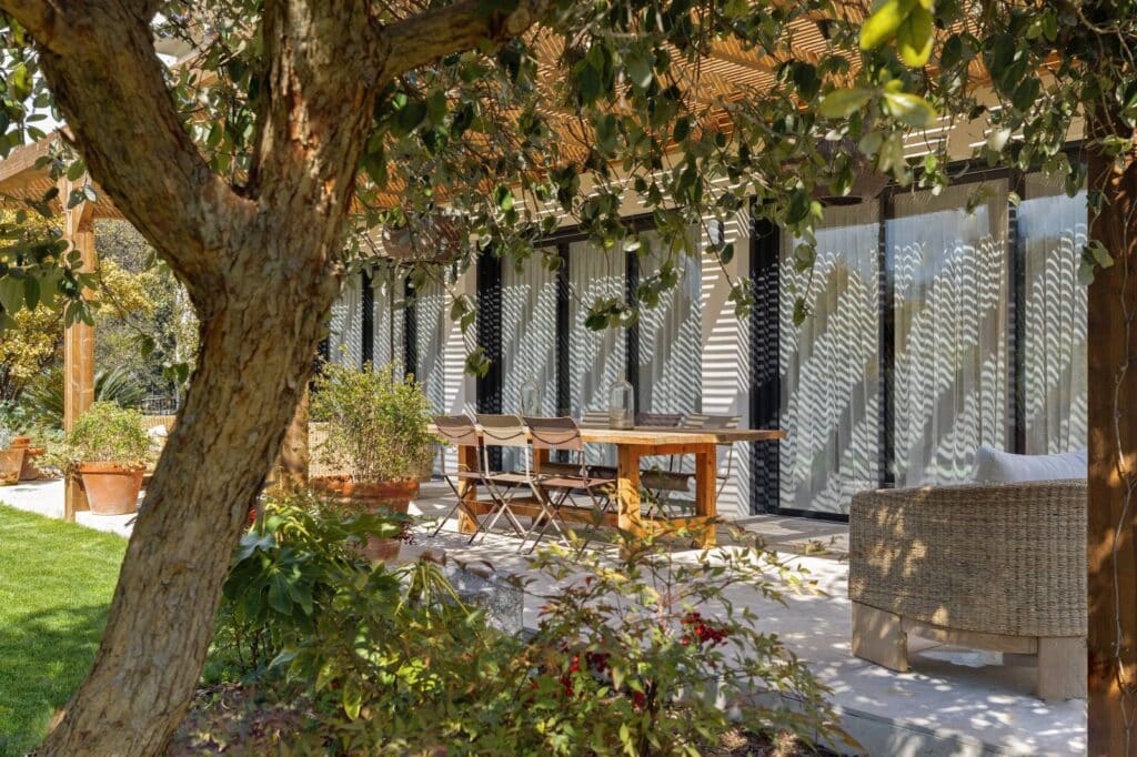 Patio ensoleillé sous pergola en bois, avec table, chaises et canapé. Un arbre projette des ombres sur la façade vitrée de la maison.