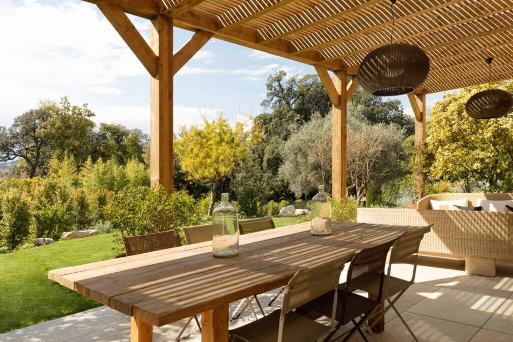 Pergola bois sur terrasse ombragée avec grande table à manger en bois, chaises et canapé en osier. Vue sur jardin luxuriant et arbres.