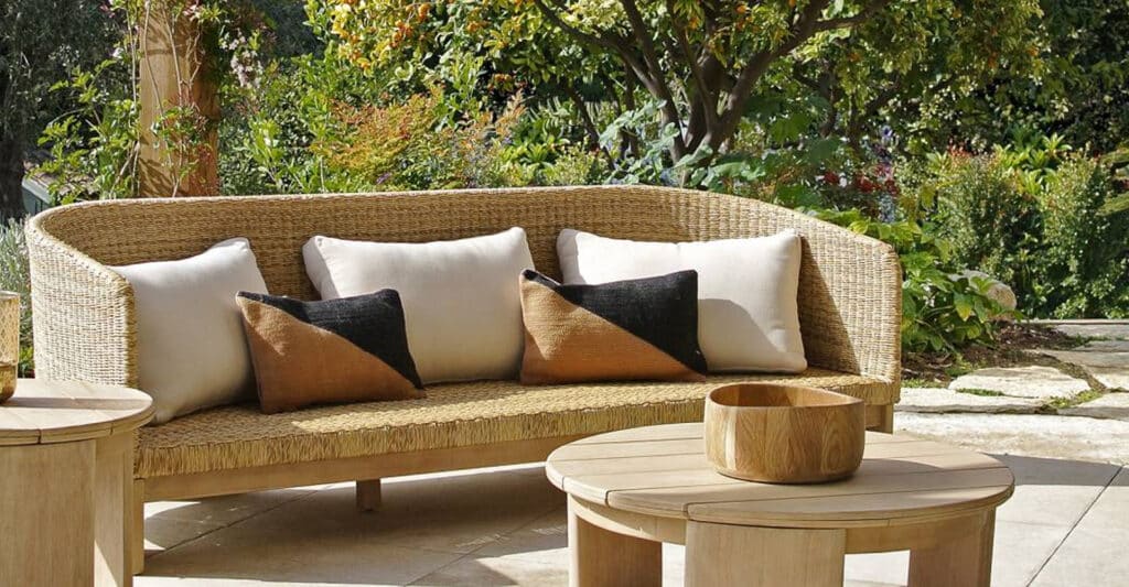 Canapé de jardin en osier clair et bois avec coussins crème et bicolores sur une terrasse ensoleillée, entouré d'un jardin luxuriant.