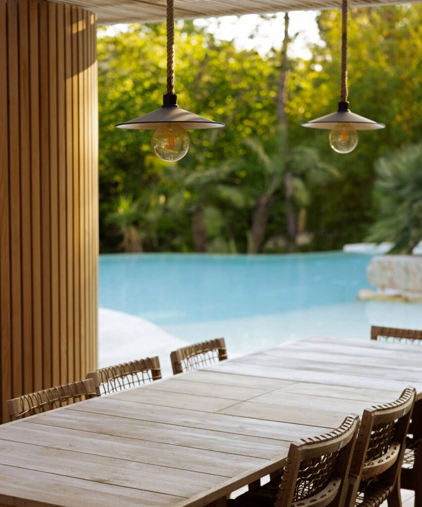 Grande table en bois clair et chaises tressées sous des suspensions rustiques près d'une piscine bleue luxuriante.