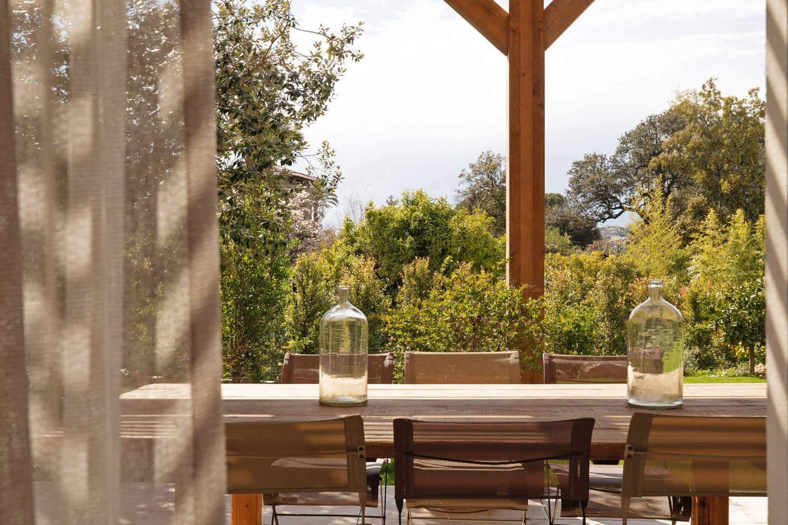 Table à manger extérieure en bois avec chaises et bouteilles, vue sur un jardin luxuriant et un ciel clair, rideau léger.