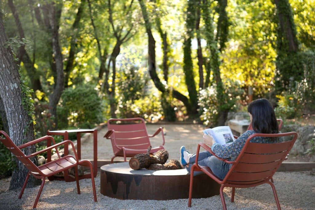 Femme lisant un magazine, détendue sur une chaise rouge près d'un brasero extérieur avec des bûches, dans un cadre boisé et ensoleillé.