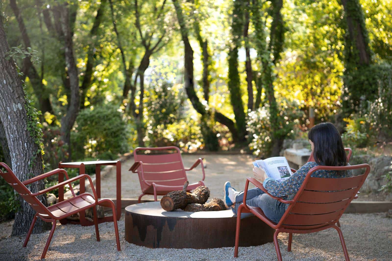 Femme lisant un magazine, détendue sur une chaise rouge près d'un brasero extérieur avec des bûches, dans un cadre boisé et ensoleillé.