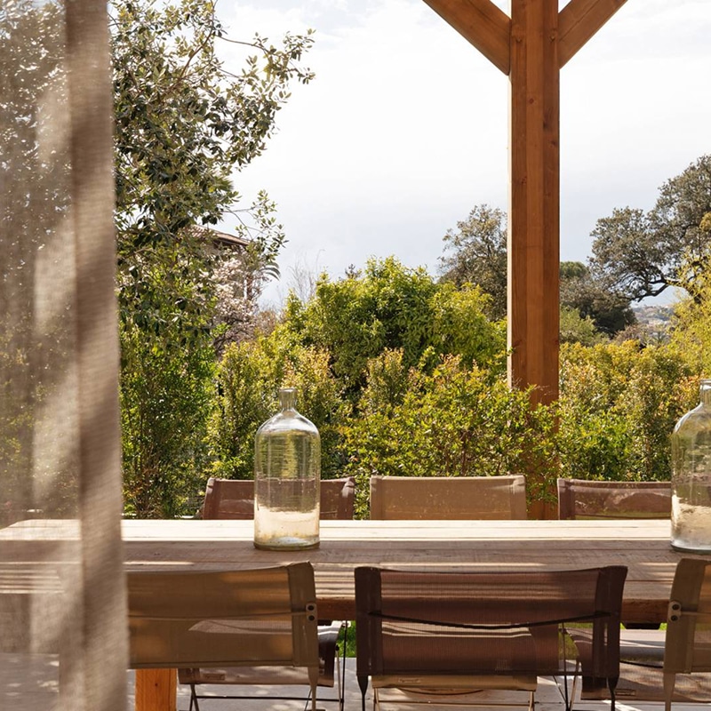 Terrasse extérieure en bois avec table, chaises beiges et bouteilles de verre, sous une pergola et entourée de végétation luxuriante.