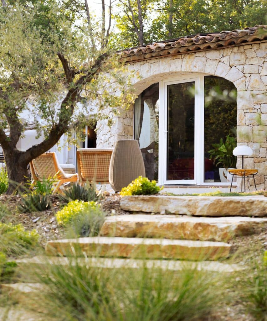 Escaliers en dalles de pierre naturelle menant à une façade en pierre sous un olivier, avec mobilier d'extérieur en rotin.