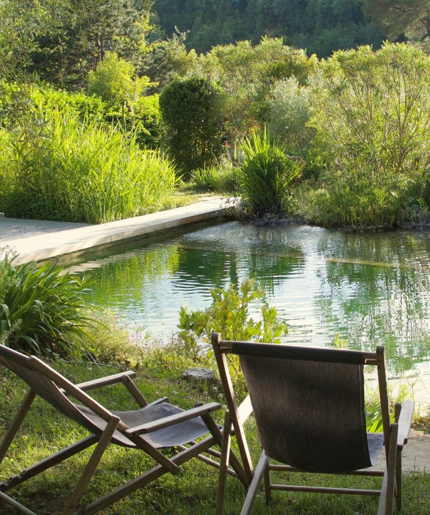 Deux chaises longues en bois font face à un bain naturel ou étang de baignade entouré de végétation luxuriante et d'herbes hautes.