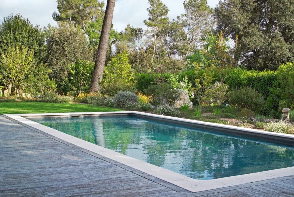 Piscine rectangulaire aux eaux bleues entourée d'un ponton en bois gris et d'un jardin luxuriant avec grands arbres et fleurs.
