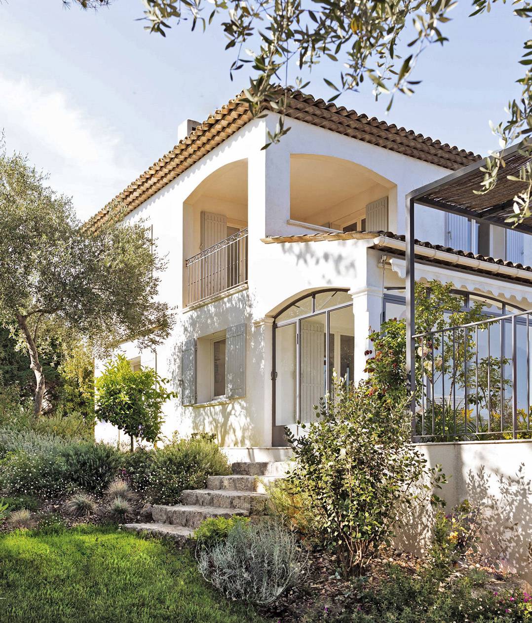 Maison blanche de style méditerranéen avec toit en tuiles, accès par escalier en pierre, entourée d'un jardin luxuriant sous un ciel bleu.