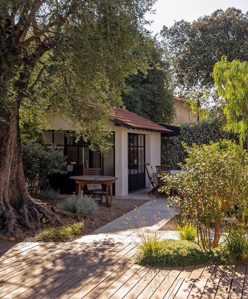 Cabane de jardin moderne avec toit tuilé rouge, terrasse en bois et mobilier d'extérieur sous un grand olivier ombragé.