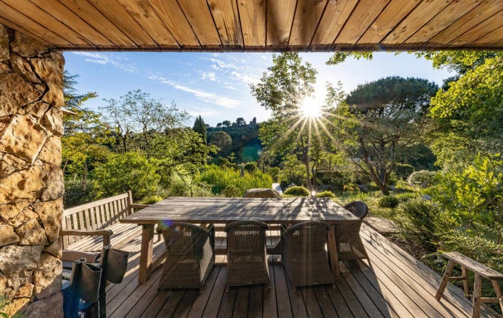 Terrasse en bois sous un toit rustique, face à un jardin verdoyant baigné de soleil avec table et chaises en osier.