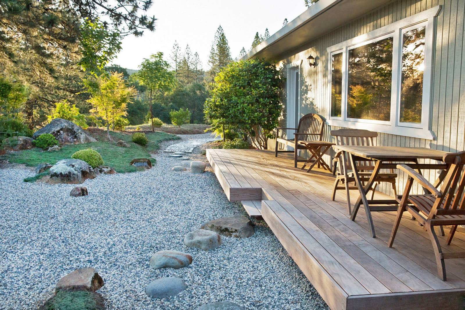 Terrasse en bois jouxtant un jardin zen avec gravier clair, rochers et végétation luxuriante au coucher du soleil.