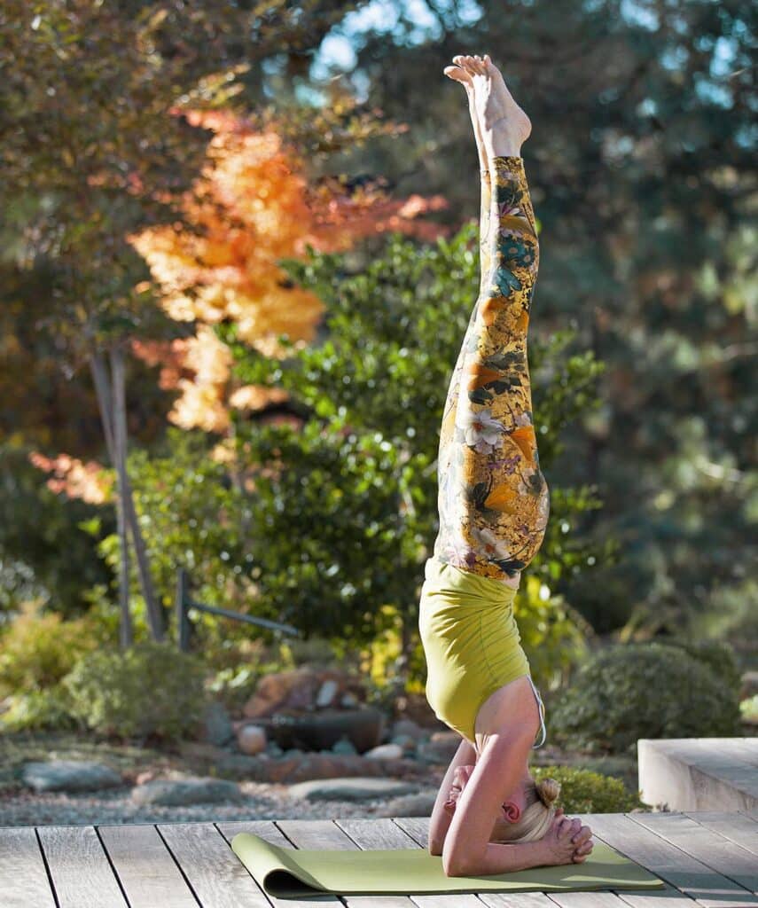 Yogi blonde en équilibre sur la tête (Sirsasana) sur un tapis vert, jambes tendues, vêtue d'un legging fleuri jaune.