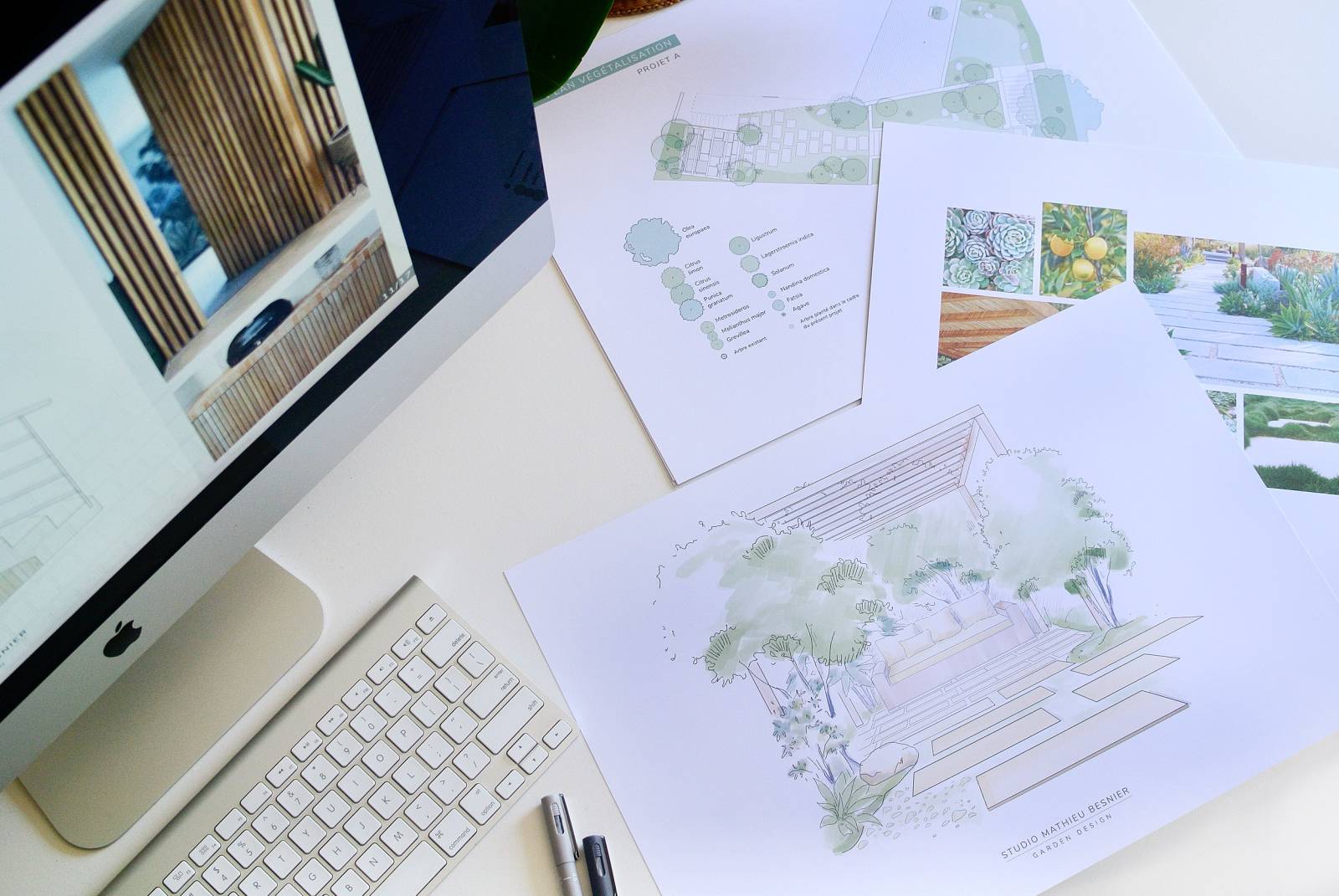 Vue en plongée d'un bureau avec un écran iMac, un clavier, des stylos et des plans de design de jardin (Projet A, Studio Mathieu Besnier) comportant croquis et végétalisation.
