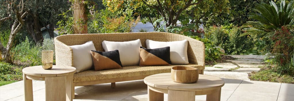 Élégant canapé en rotin tressé avec coussins clairs et tables d'appoint en bois sur une terrasse ensoleillée, entouré d'un jardin luxuriant.