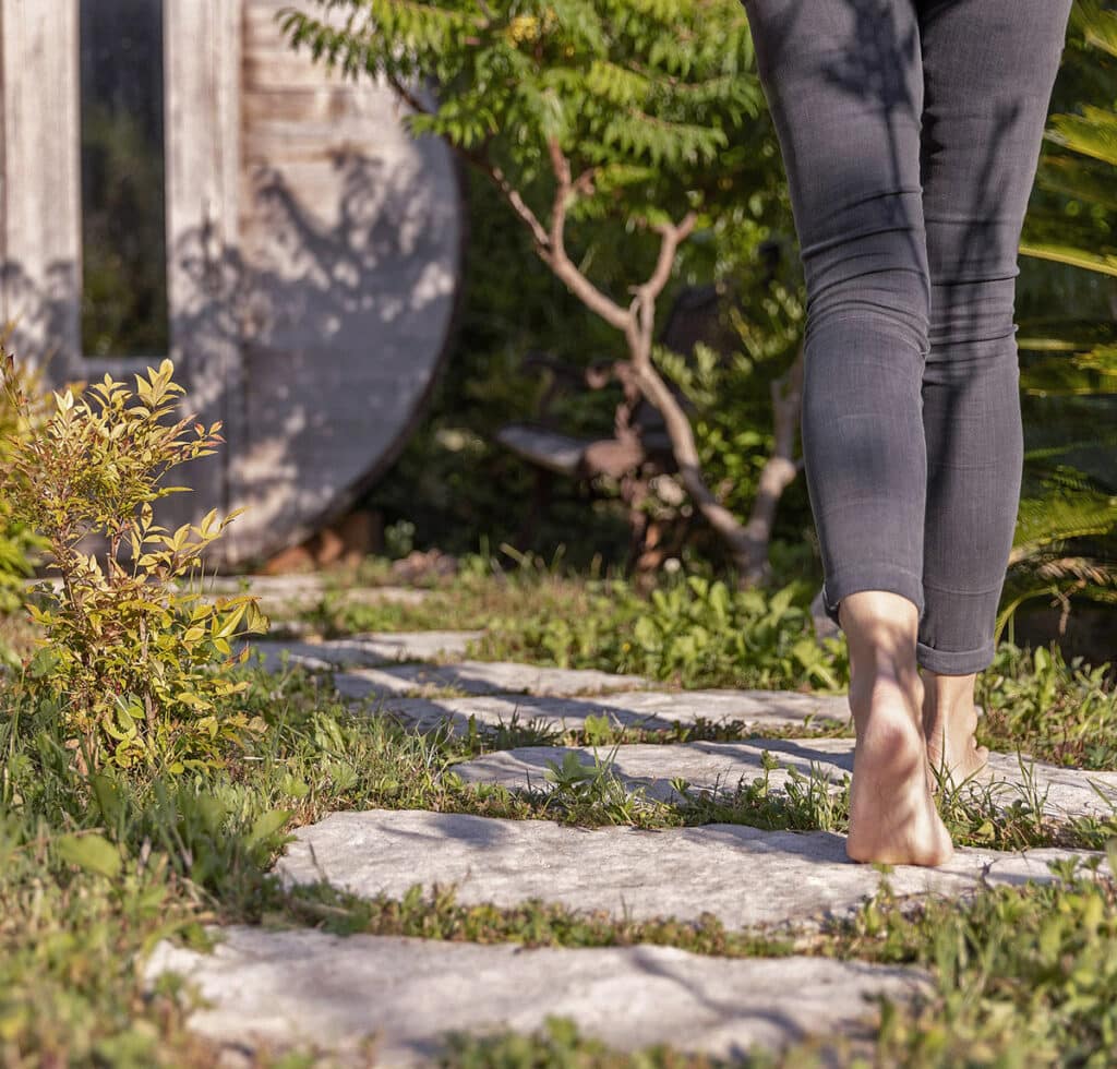 Jambes et pieds nus marchant sur un sentier de dalles de pierre traversant l'herbe haute d'un jardin ensoleillé.