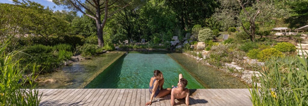 Couple relaxé sur une terrasse en bois au bord d'un bassin de nage naturel et écologique, entouré d'une végétation luxuriante.
