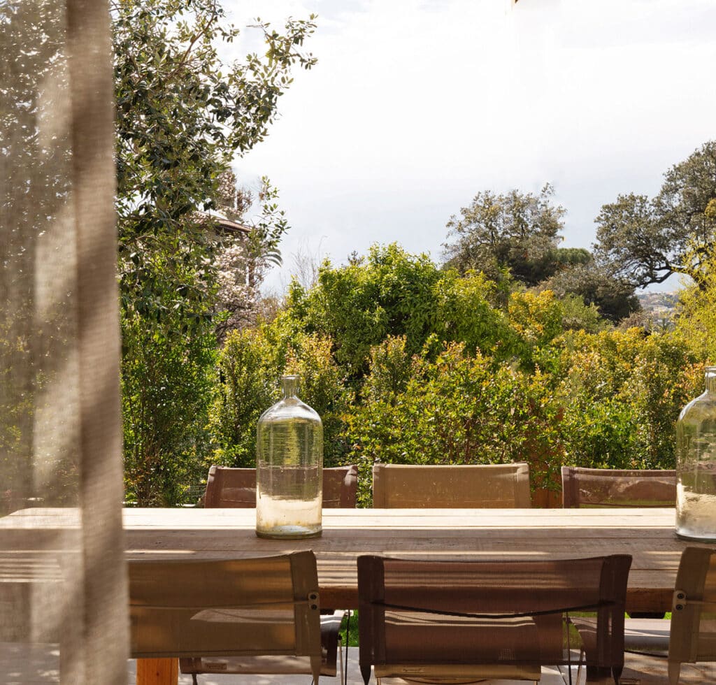 Table en bois sur une terrasse abritée, avec chaises et deux bouteilles, offrant une vue dégagée sur un jardin luxuriant.