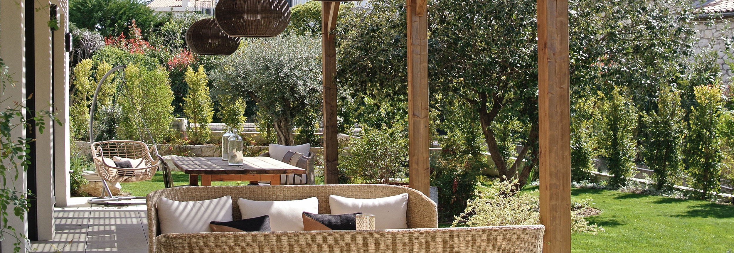 Patio ensoleillé, canapé en rotin et table en bois sous pergola. Grand jardin verdoyant avec olivier, ambiance méditerranéenne.