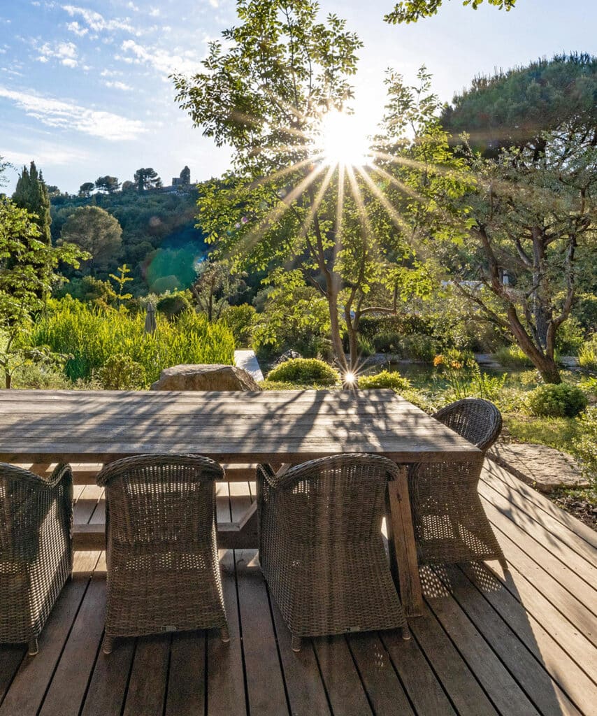 Table et chaises en osier sur un deck en bois. Ambiance jardin luxuriant, soleil éclatant à travers les arbres et ruines sur la colline.